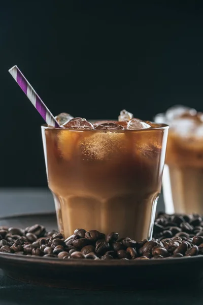 Selective focus of glass of cold iced coffee with straw on plate with roasted coffee beans on tabletop on dark backdrop — Stock Photo