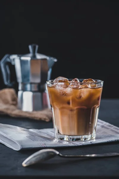 Selective focus of glass of cold iced coffee, newspaper and coffee maker on tabletop on dark backdrop — Stock Photo