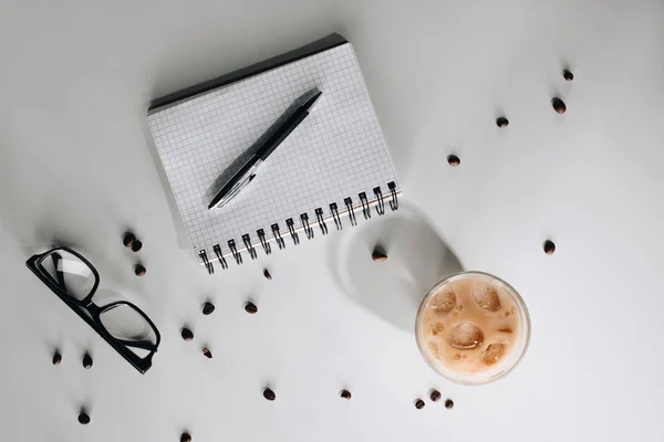 Flache Liege mit Glas kalten Eiskaffees, gerösteten Kaffeebohnen, Brille, leerem Notizbuch und Stift auf weißer Oberfläche — Stockfoto