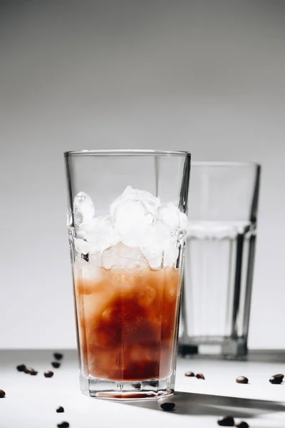 Vue rapprochée du verre de café infusé à froid avec glaçons et grains de café torréfiés sur fond gris — Photo de stock