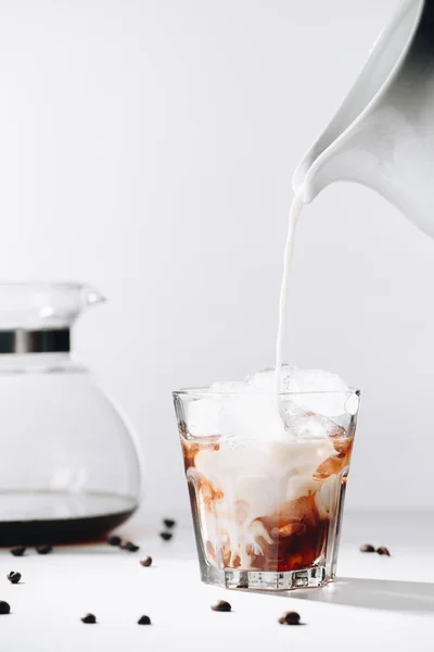 Primer plano vista de verter la leche en el vaso de proceso de café elaborado en frío, cafetera y granos de café tostados sobre fondo gris - foto de stock