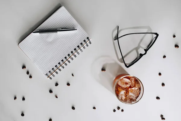 Tendido plano con vaso de café helado frío, granos de café tostados, anteojos, cuaderno vacío y pluma en la superficie blanca - foto de stock