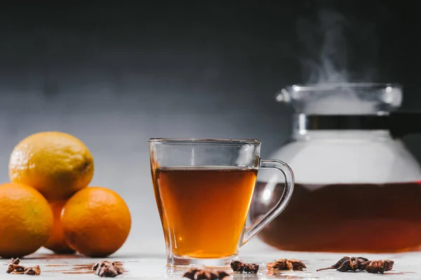 Cup of hot black tea with citrus fruits and steaming teapot — Stock Photo