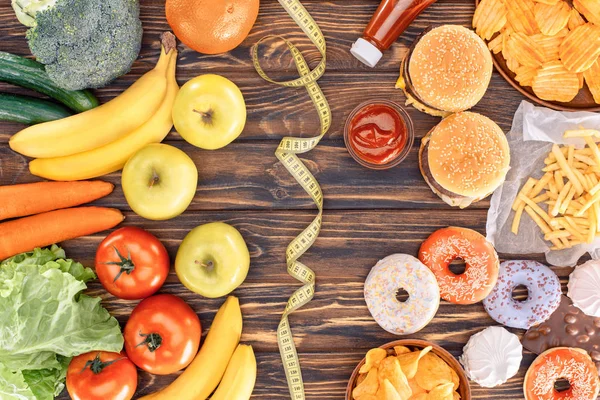 Top view of assorted junk food, fresh fruits with vegetables and measuring tape on wooden table — Stock Photo