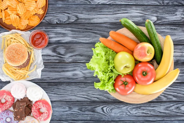 Vista superior de frutas frescas con verduras y platos con comida chatarra en mesa de madera - foto de stock