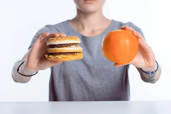 Tiro cortado de menina segurando hambúrguer e laranja madura — Fotografia de Stock