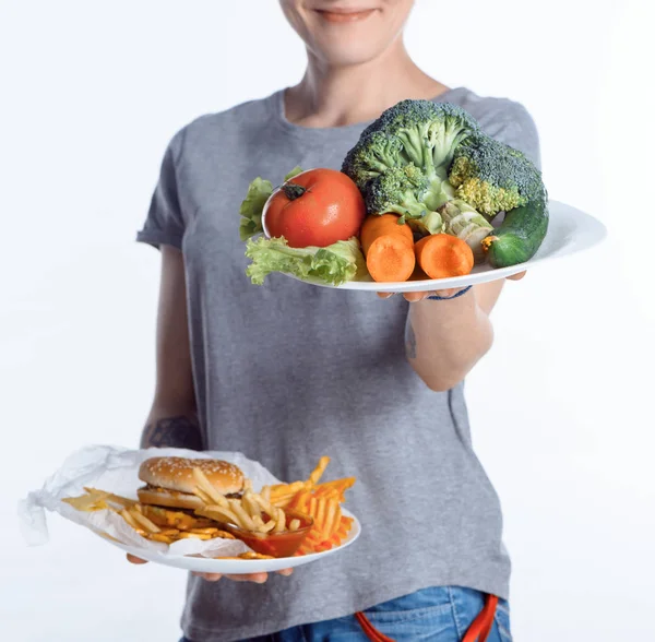 Tiro cortado de mulher segurando pratos com legumes e junk food — Fotografia de Stock