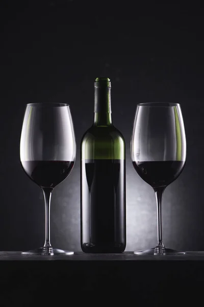 Glasses filled with red wine and wine bottle in middle on black — Stock Photo