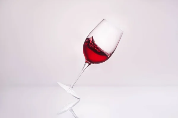 Verre à vin incliné avec éclaboussures de vin rouge sur la surface réfléchissante et sur blanc — Photo de stock
