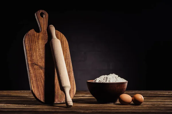 Closeup view of wooden cutting board, rolling pin, bowl with flour and two eggs on table — Stock Photo