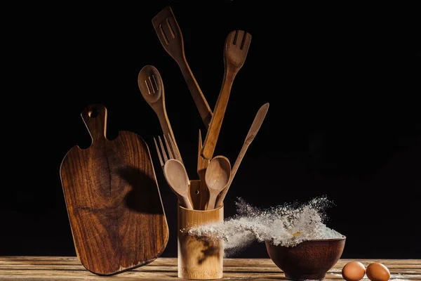 Flying cutting board, kitchen utensils, flour from bowl and two eggs on table on black background — Stock Photo