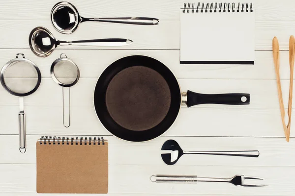 Vista superior de libros de texto, sartén y utensilios de cocina sobre mesa de madera blanca - foto de stock