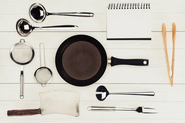 Vista dall'alto del libro di testo in bianco, padella, ascia macellaio e utensili da cucina su tavolo di legno bianco — Foto stock