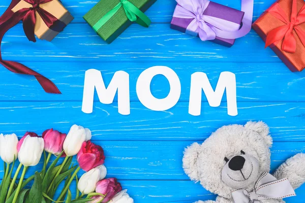 Vista superior de cajas de regalo, oso de peluche, tulipanes y palabra mamá en la mesa azul, concepto de día de las madres - foto de stock