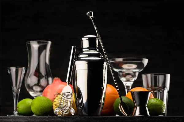 Shaker brilhante para preparar bebida alcoólica, copos vazios e frutas maduras na mesa isolada em preto — Fotografia de Stock