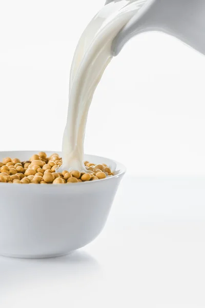 Leche vertiendo de la jarra en un tazón con soja sobre fondo blanco - foto de stock