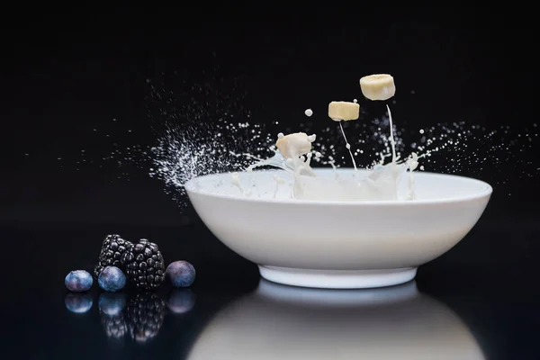 Banana y bayas cayendo en tazón blanco con leche sobre fondo negro - foto de stock