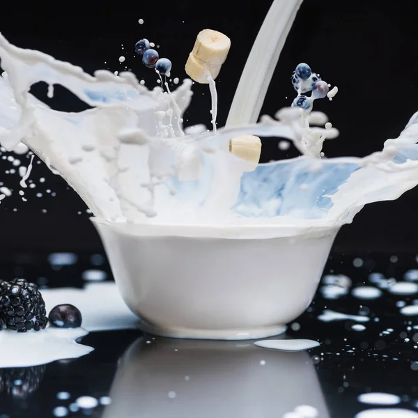 Splashing milk with banana and berries in white bowl on black background — Stock Photo