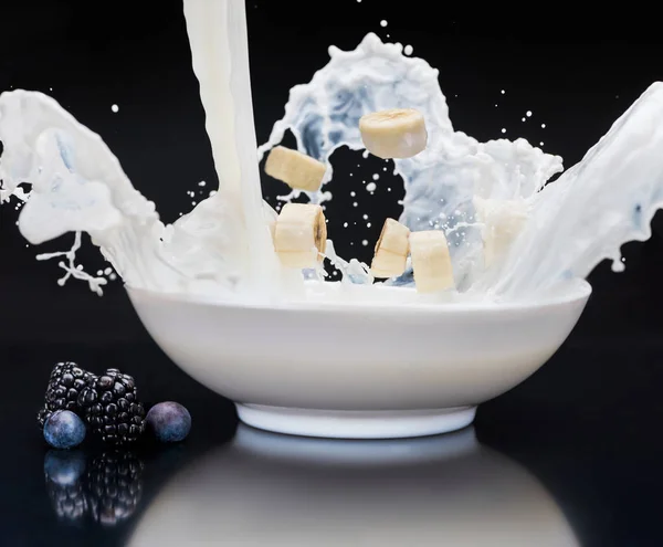 Bananenstücke fallen in weiße Schüssel mit Milch auf schwarzem Hintergrund — Stockfoto