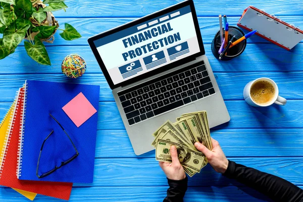 Geschäftsmann zählt Dollars per Laptop auf blauem Holztisch mit Schreibwaren, Finanzschutz-Schriftzug — Stockfoto