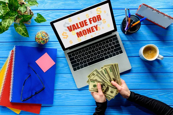 Geschäftsmann zählt Dollar per Laptop auf blauem Holztisch mit Schreibwaren, Preis-Leistungs-Verhältnis Schriftzug — Stockfoto