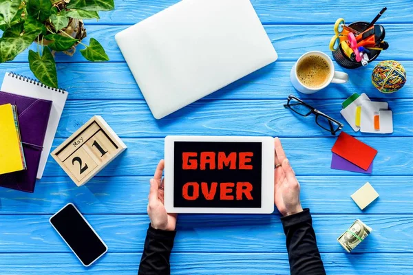 Hands holding tablet on blue wooden table with money and stationery, Game over lettering — Stock Photo