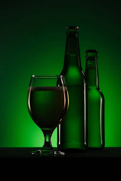 Vista de cerca de botellas y vaso de cerveza sobre fondo verde oscuro - foto de stock