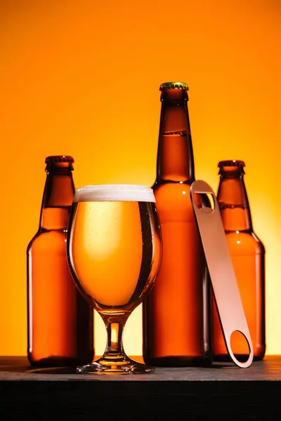 Vista de cerca de botellas, vaso de cerveza con espuma y abridor de botellas sobre fondo naranja - foto de stock