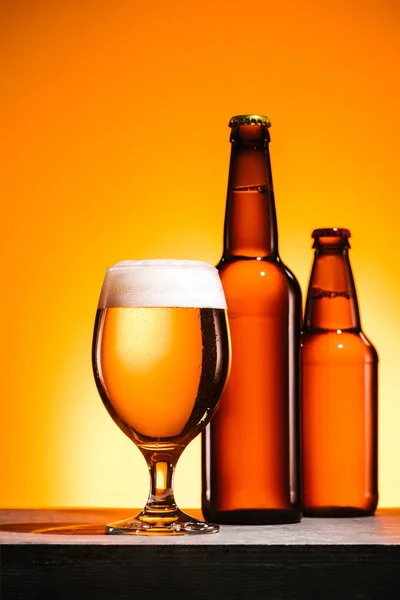 Close up view of bottles and glass of beer with foam on surface on orange background — Stock Photo