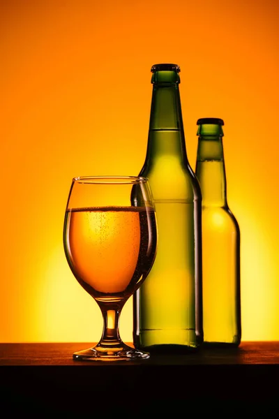 Vista de cerca de botellas y vaso de cerveza en la superficie sobre fondo naranja - foto de stock