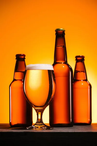 Close up view of bottles and glass of beer with foam on surface on orange background — Stock Photo