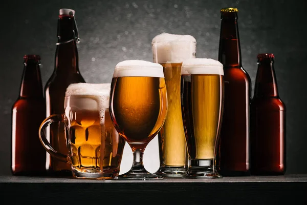 Закрыть вид бутылок и кружек пива с пеной на сером фоне — стоковое фото