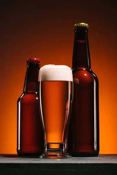 Close up view of bottles and mug of beer with foam on orange backdrop — Stock Photo