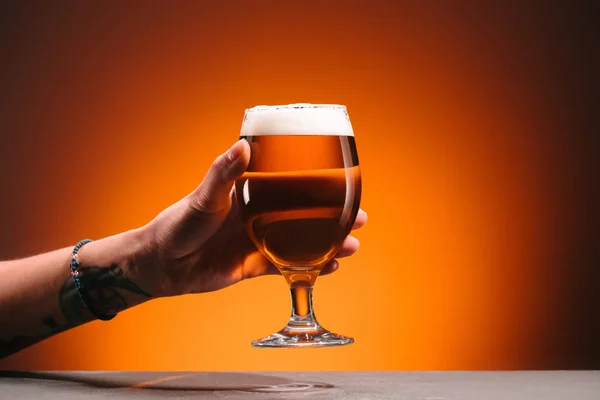 Tiro recortado de mulher segurando vidro de cerveja fria com espuma no fundo laranja — Fotografia de Stock