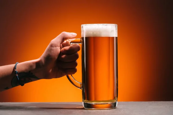 Tiro recortado de mujer sosteniendo vaso de cerveza fría con espuma sobre fondo naranja - foto de stock