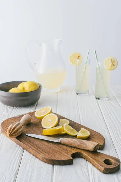 Close up view of lemons, knife, wooden pestle on cutting board and lemonade in glasses on white wooden tabletop on grey backdrop — Stock Photo