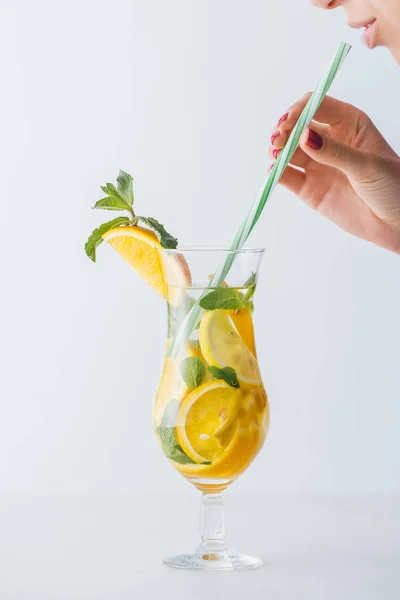 Partial view of woman holding straw and summer cold cocktail with mint, lemon and orange pieces isolated on white — Stock Photo