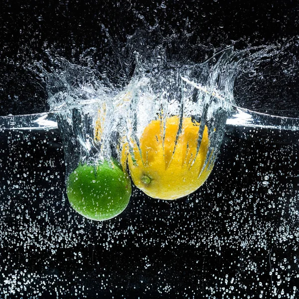 Vue rapprochée des citrons frais et des citrons verts dans l'eau isolée sur noir — Photo de stock