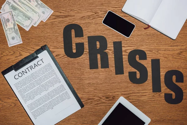 Top view of word crisis near smartphone, digital tablet, notebook, contract on clipboard and dollar banknotes on wooden desk — Stock Photo