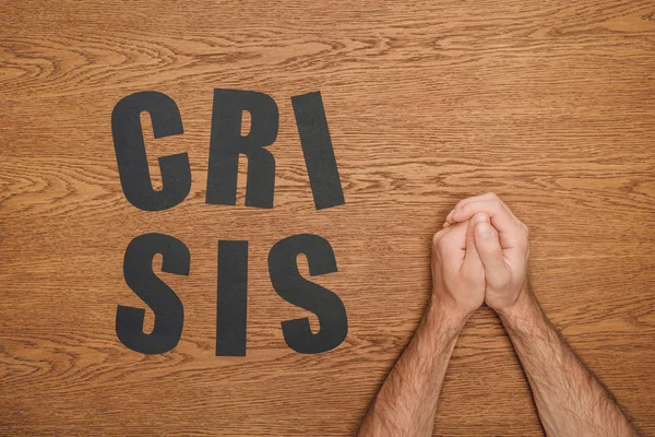 Cropped view of male folded hands near black paper cut word crisis on wooden desk — Stock Photo