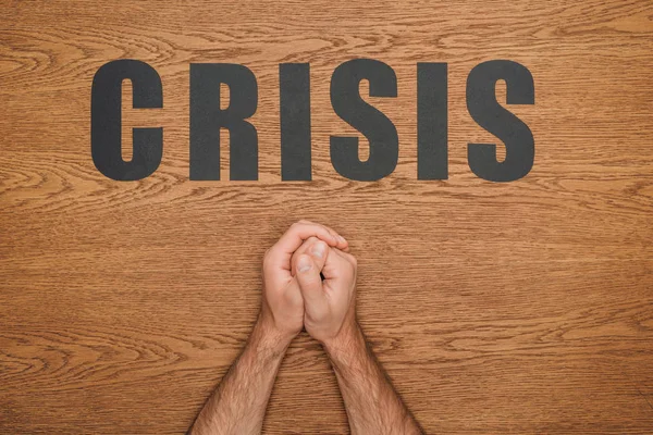 Cropped view of male folded hands near black paper cut word crisis on wooden desk — Stock Photo