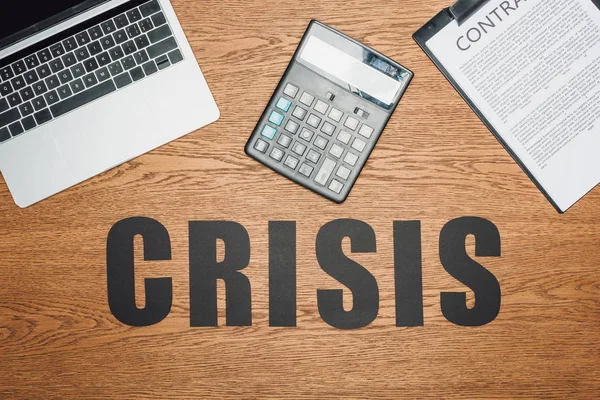 Top view of black word crisis near laptop, calculator and clipboard with contract on wooden desk — Stock Photo