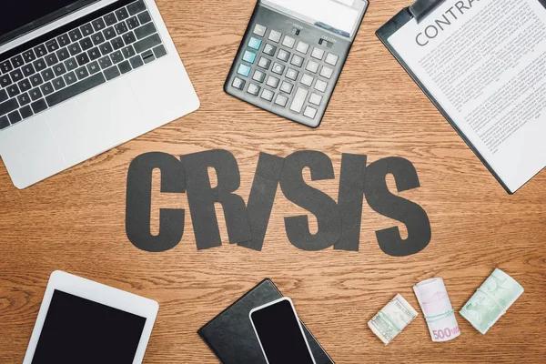 Top view of word crisis near digital devices, clipboard with contract, cash rolls of dollar, euro and ruble banknotes on wooden desk — Stock Photo