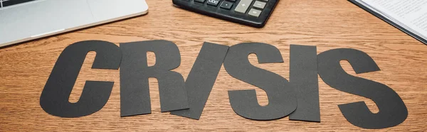 Panoramic shot of black paper cut word crisis near laptop, calculator and document on wooden desk — Stock Photo