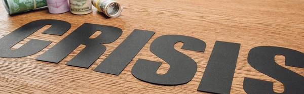 Panoramic shot of black paper cut word crisis near cash rolls on wooden surface — Stock Photo