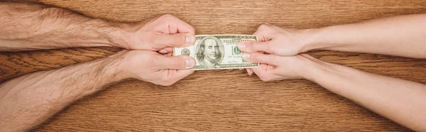 Partial view of man and woman holding dollar banknote on wooden surface, panoramic shot — Stock Photo