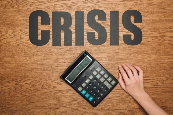 Vista recortada de la mano femenina cerca de la calculadora con cien mil en pantalla, y la palabra crisis en la superficie de madera - foto de stock