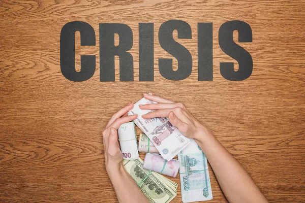 Cropped view of woman grabbing packs of dollar, euro and ruble banknotes, and paper cut word crisis on wooden desk — Stock Photo