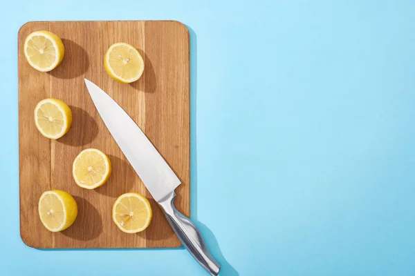 Top view of ripe yellow cut lemons on wooden cutting board with knife on blue background with copy space — Stock Photo