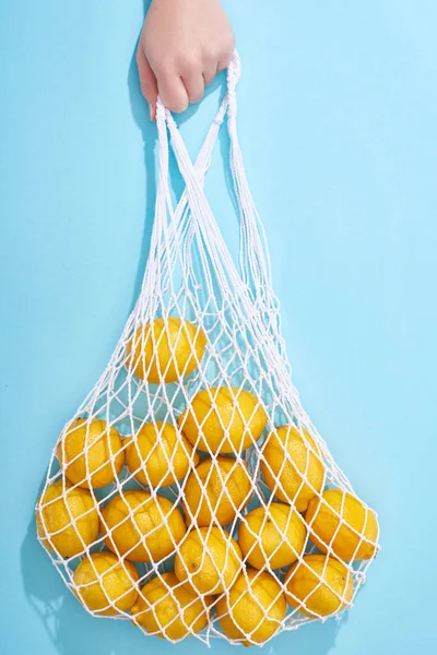 Vista recortada de mujer sosteniendo limones amarillos maduros en bolsa de hilo sobre fondo azul - foto de stock